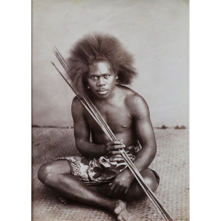 PHOTOGRAPHER UNKNOWN [attrib. to Thomas Andrews 1855-1939] - Portrait of a young Samoan man sitting on a flax mat holding spears
